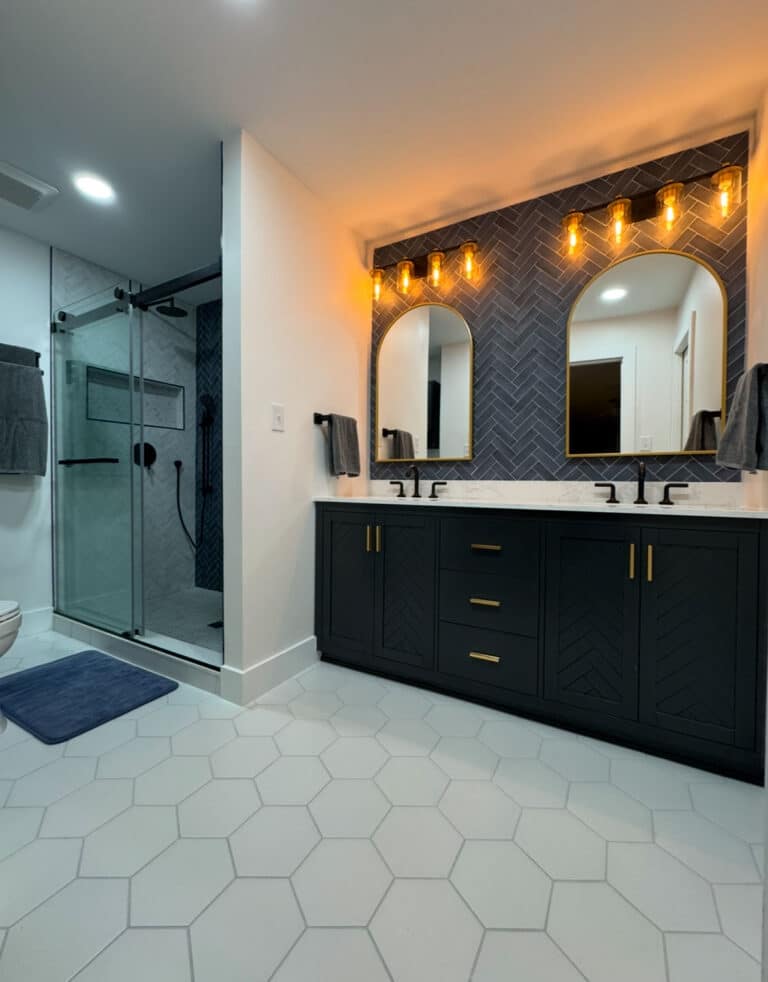 Contemporary bathroom with dual-sink vanity, black cabinetry, gold hardware, arched mirrors, exposed bulb lighting, walk-in shower with sliding glass doors, and hexagonal floor tiles.