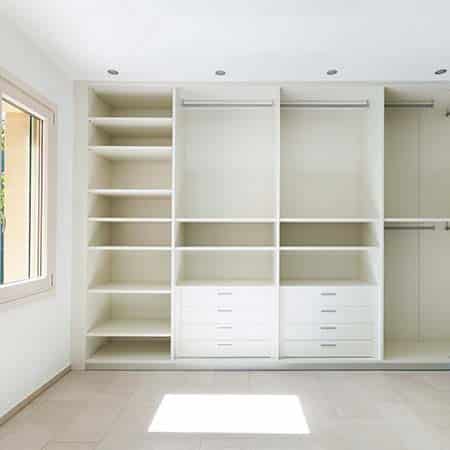 Empty built-in white closet system with open shelves, drawers, and hanging rods, situated in a well-lit room with a window on the left.
