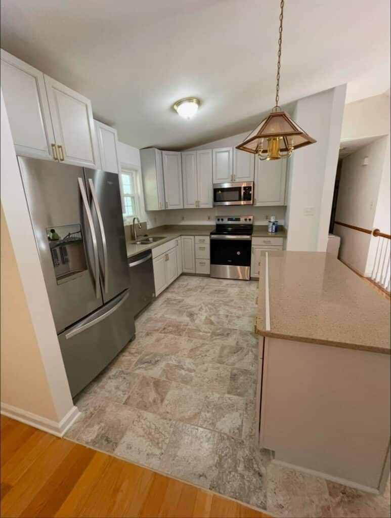 Modern kitchen with white cabinetry, stainless steel appliances, beige stone tile flooring, a quartz countertop island, and pendant lighting over the counter.