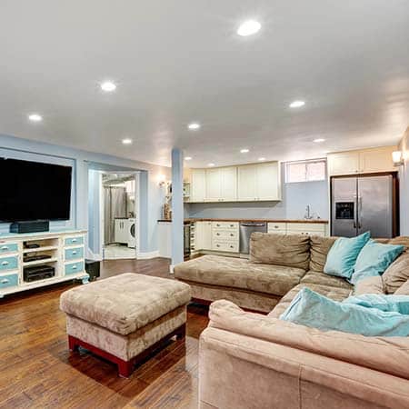 Finished basement with wood flooring, a large sectional sofa, flat-screen TV, and an open kitchenette with white cabinets and stainless steel appliances.