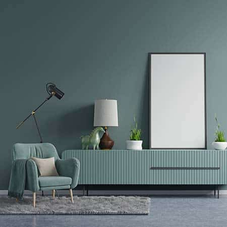 Modern living space with a muted green wall, matching armchair, floor lamp, sideboard with decorative items, and a large blank frame leaning against the wall.