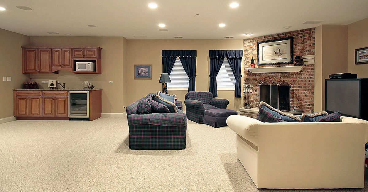 Finished basement featuring a cozy living area with plaid upholstered furniture, a brick fireplace with framed art above the mantel, and a small kitchenette with wooden cabinets, a microwave, and a mini fridge. Two windows with navy curtains provide natural light.