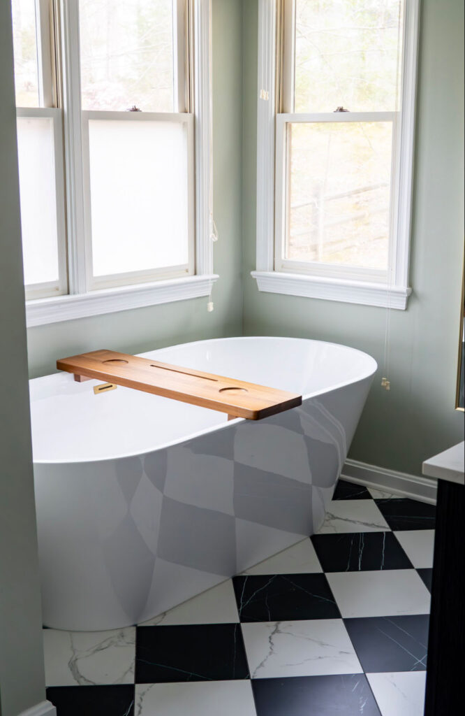 Freestanding white bathtub with wooden tray, positioned by large windows, set on a black and white checkered tile floor in a light green bathroom.
