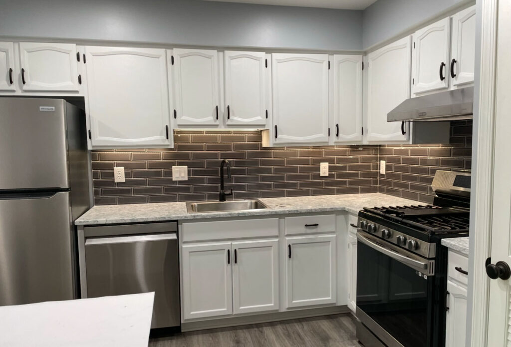 Remodeled kitchen with white cabinets, stainless steel appliances, black subway tile backsplash, gray countertops, and under-cabinet lighting.
