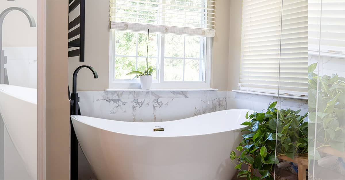 Freestanding white bathtub under a window with blinds, surrounded by indoor plants.