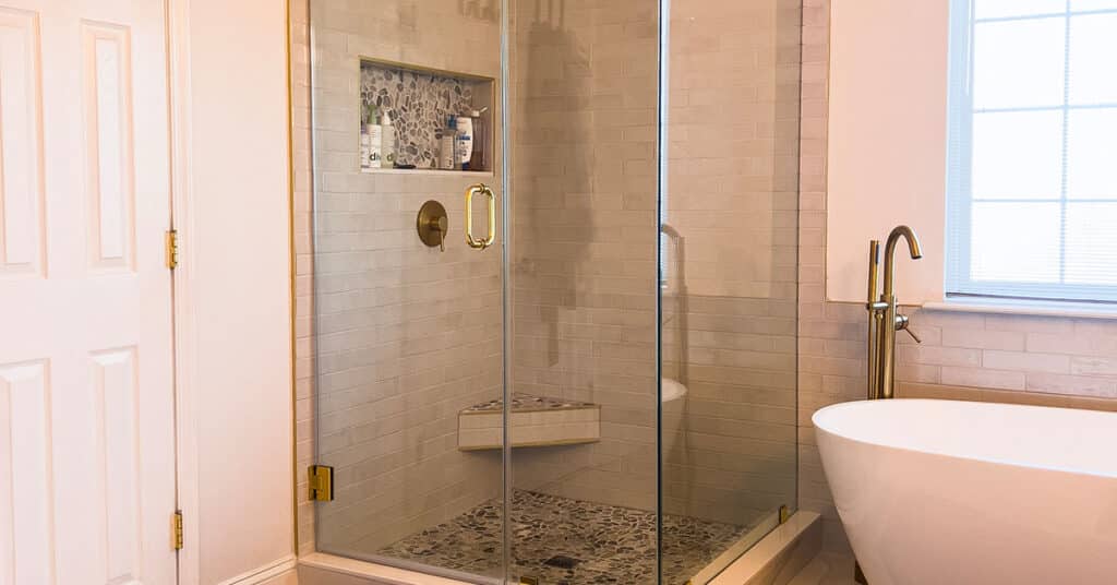 Glass shower with gold hardware, niche and bench; pebble floor beside freestanding tub.