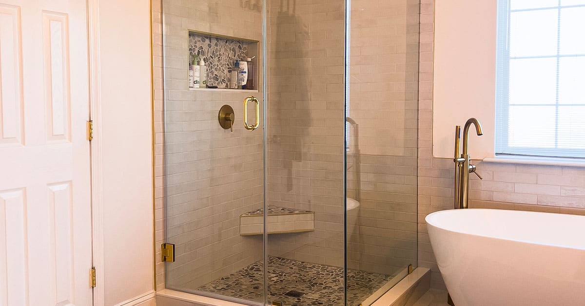 Glass shower with gold hardware, niche and bench; pebble floor beside freestanding tub.